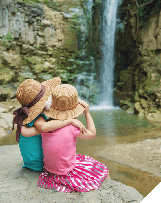 Viajes en familia. Experiencias diferentes. Aventura. Dos niñas con sombrero de paja miran una cascada de agua