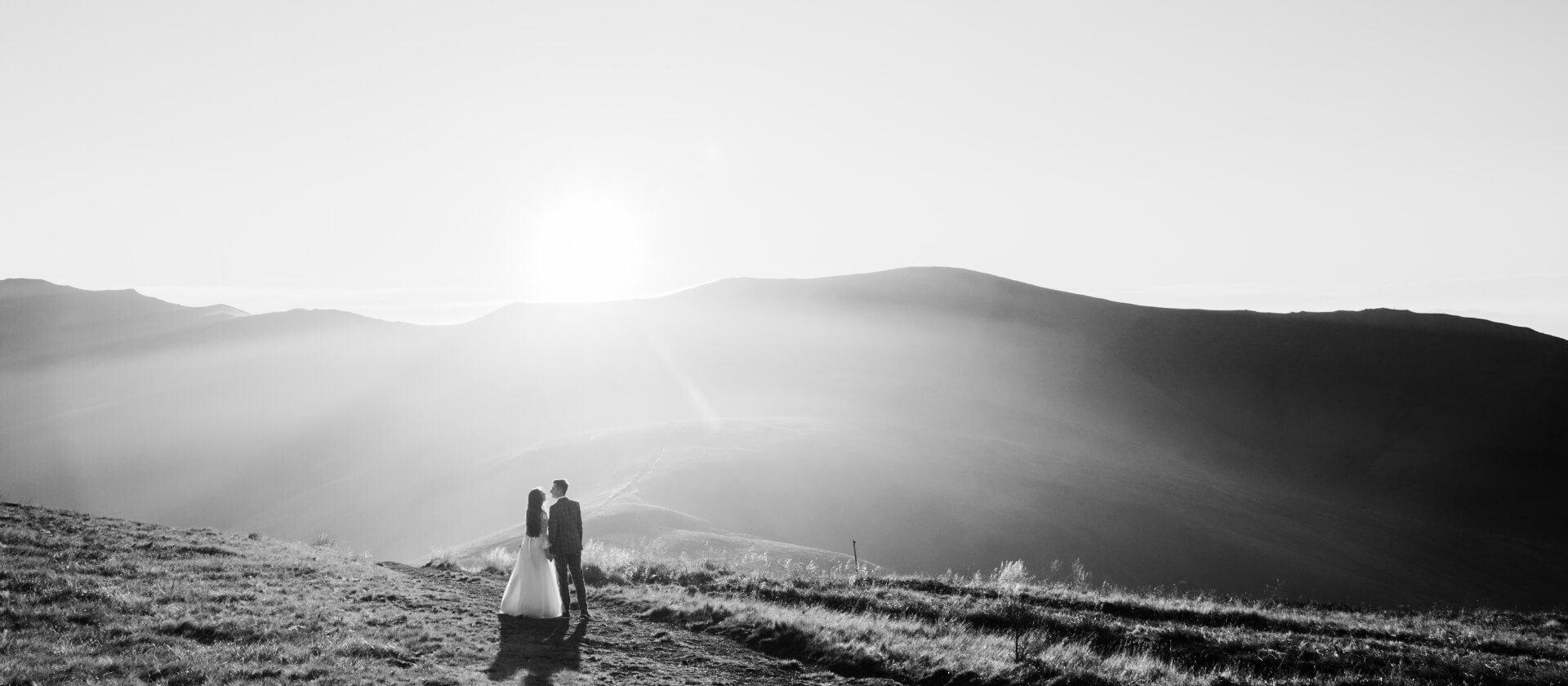Viajes de novios. Una pareja vestidos de ceremonia en la montaña miran al horizonte en la puesta del sol