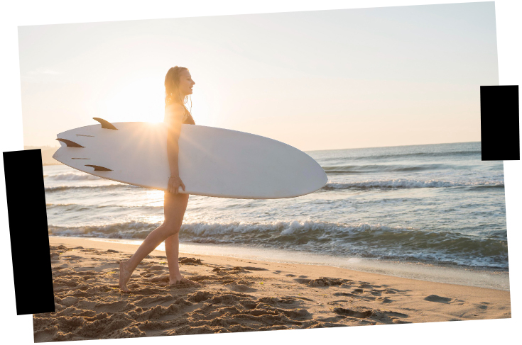 Viajes a medida. Viajes que inspiran. Una mujer con su tabla de surf camina hacia el agua en la orilla del mar al atardecer