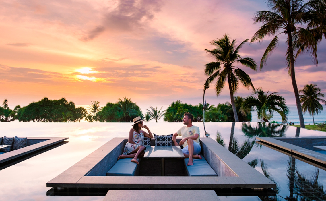 Hombre y mujer contemplando el atardecer desde una piscina infinity en un exclusivo resort de Tailandia.